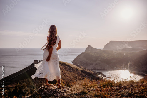 A woman in a white dress stands on a hill overlooking the ocean. The scene is serene and peaceful, with the woman's dress billowing in the wind. The combination of the ocean.