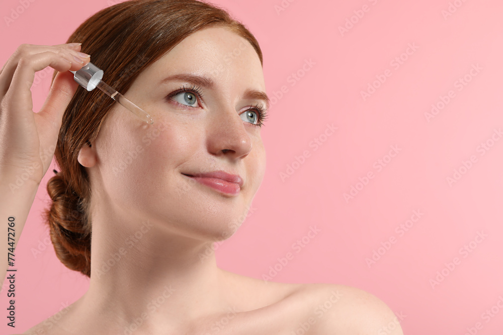 Beautiful woman with freckles applying cosmetic serum onto her face on pink background, closeup. Space for text