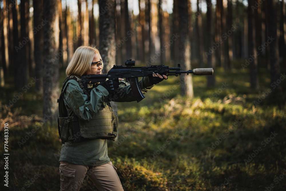 Blonde young military girl aims with a modern AK 12 rifle in the forest at war. Blonde young military girl aims with a modern AK 12 rifle in the forest at war.