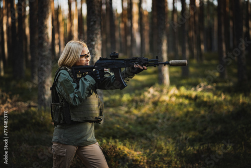 Blonde young military girl aims with a modern AK 12 rifle in the forest at war.