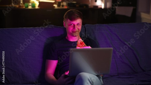 Portrait of cheerful young man watching content on laptop screen and eating pizza at night sitting alone on sofa. Happy male watching movie or online football game, relaxing after workday, slow motion photo