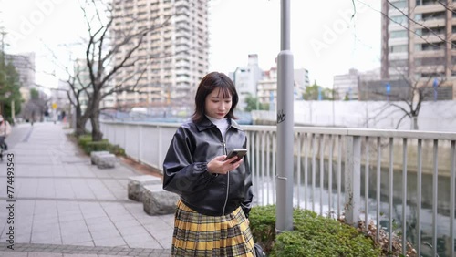 A Japanese woman in her 20s walking along the riverside road around Gotanda Station, Shinagawa-ku, Tokyo in winter while operating her smartphone 冬の東京都品川区五反田の駅周辺の川沿いの道をスマートフォンを操作しながら歩く２０代の日本人女性  photo