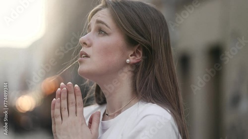 Portrait of hopeful young woman clasping hands in prayer asking for blessing and help while the rays of rising sun fall on her face at city outdoors Religion and faith concept photo