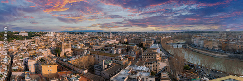 An aerial view of Rome at dusk reveals a vibrant sky in orange, pink, purple, and blue. Ancient and modern buildings, terracotta roofs, and church domes stand out. The Tiber River enhances the scene. photo