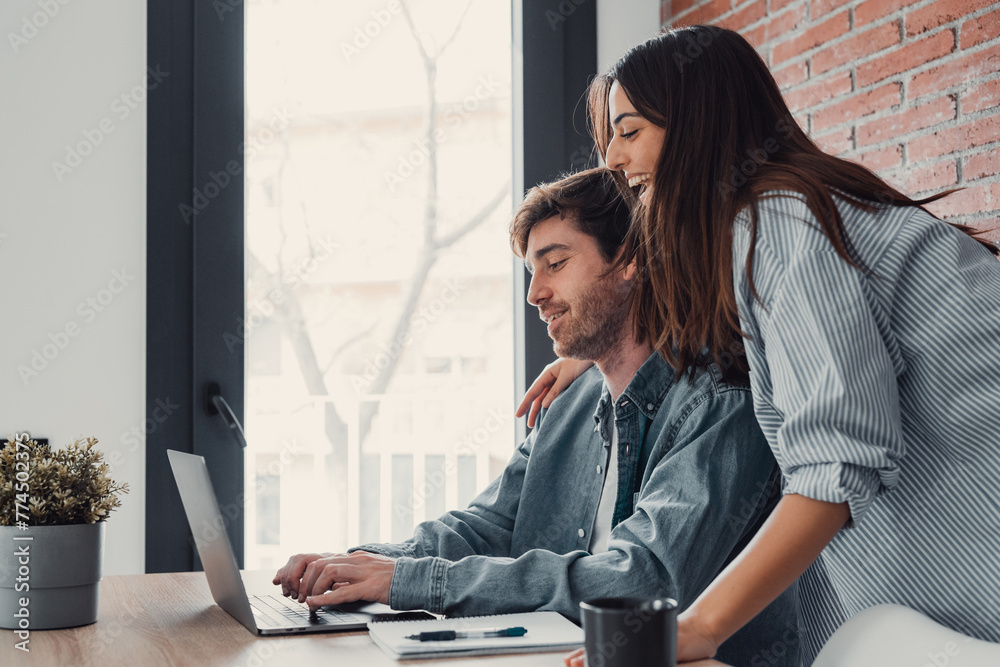 Naklejka premium Happy young family couple bonding laughing using laptop looking at screen sit at table, smiling husband and wife buying choosing goods online doing internet shopping order with delivery at home.
