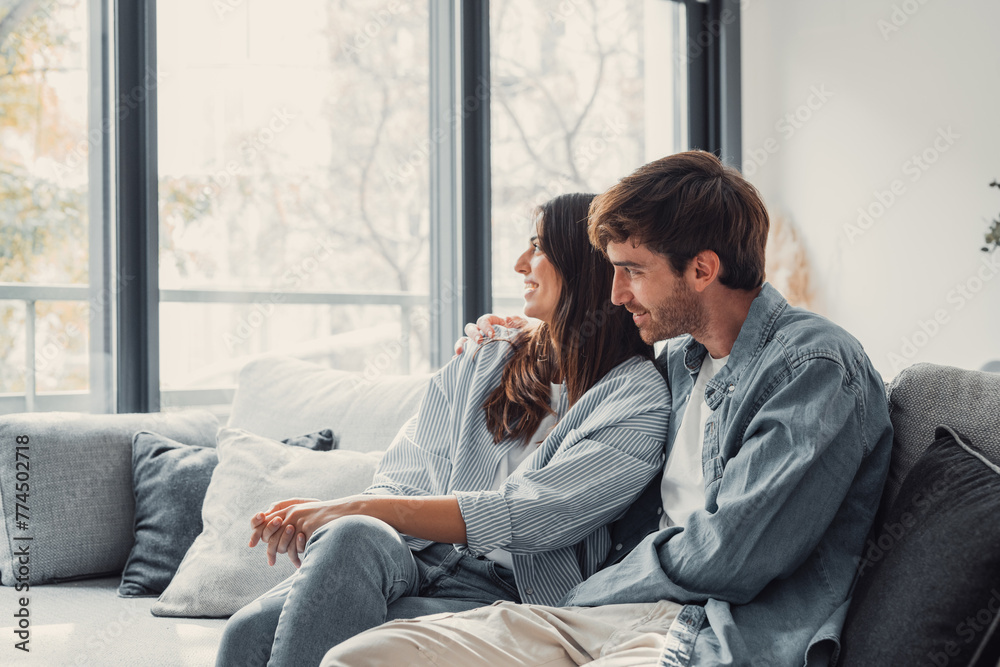 One dream for two of us. Romantic millennial spouses relax on couch together looking aside imagining happy future life. Young man and woman in love cuddling indoors creating plans on weekend vacation.