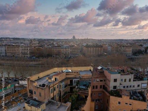 Rome from above at dawn or dusk, with terracotta roofs and the winding Tiber River. St. Peter's Basilica's dome stands out against a pink and blue sky. photo