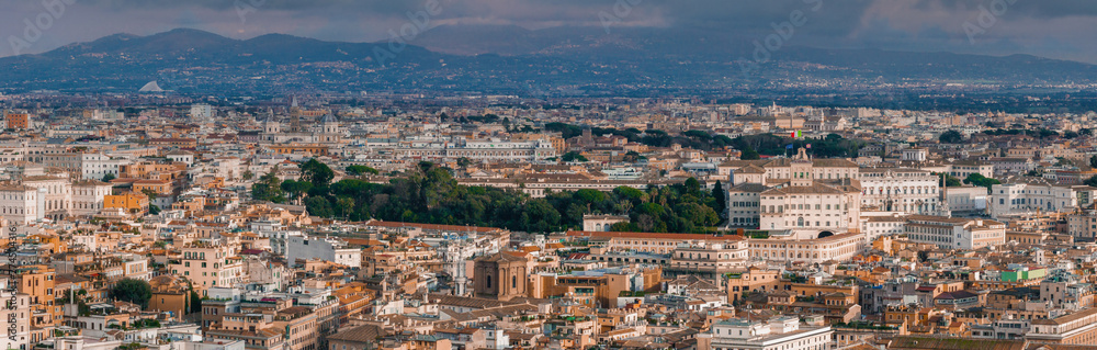 An aerial view of Rome shows a dense cityscape, terracotta roofs, and mixed architecture under a cloudy sky. Its rich history is seen in the mix of old and new buildings, nestled among hills.