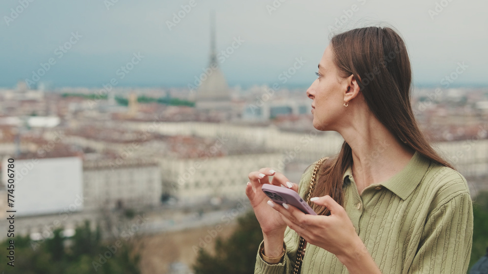 Young woman with brown hair wearing an olive green sweater on the observation deck of the city uses a mobile phone - obrazy, fototapety, plakaty 
