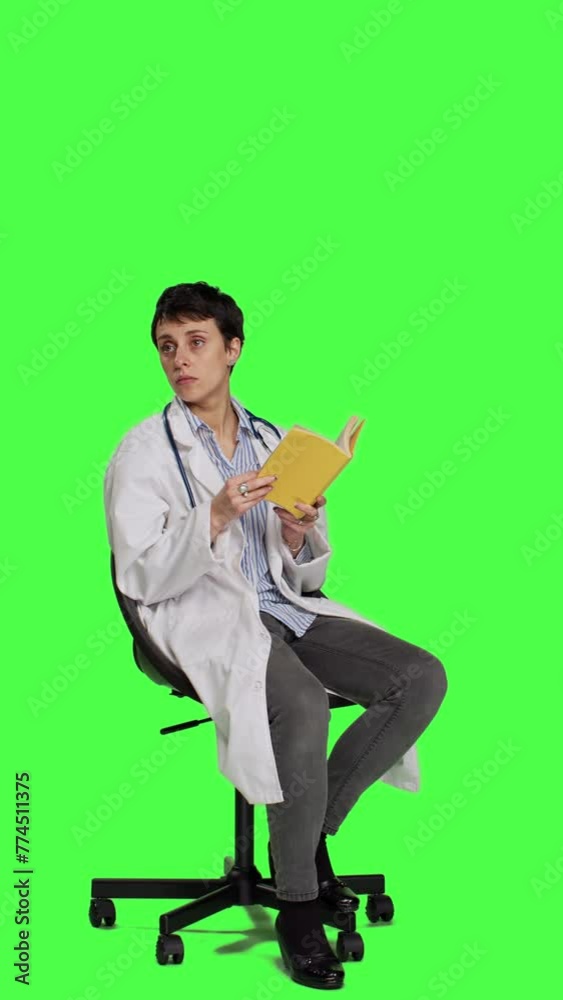 Side view Medic specialist reading a medicine literature book sitting on a chair, enjoying healthcare industry lecture against greenscreen backdrop. Doctor in white coat reads for relaxation. Camera A Side view Medic specialist reading a medicine literature book sitting on a chair, enjoying healthcare industry lecture against greenscreen backdrop. Doctor in white coat reads for relaxation. Camera A