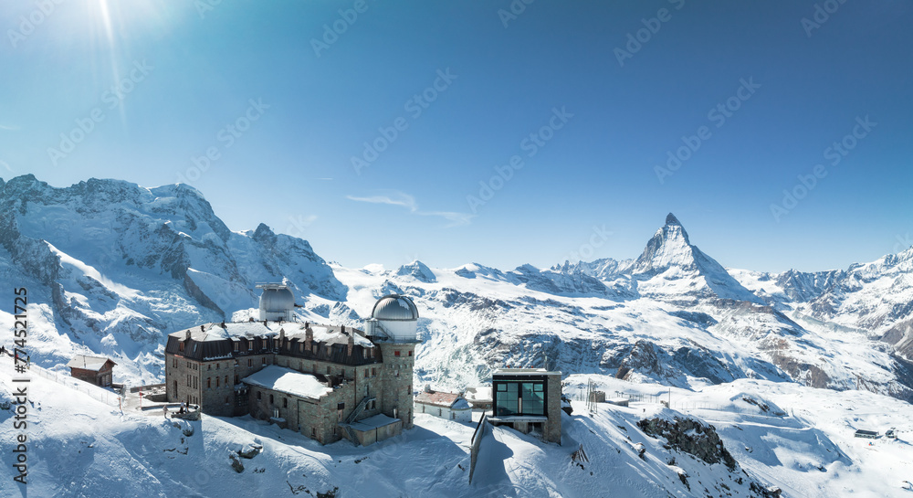 An aerial shot shows Zermatt, Switzerland's snowy scene, with a large dome building and the iconic Matterhorn peak under a clear sky, ideal for skiing and mountaineering.