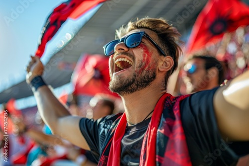 A man wearing sunglasses and a red scarf is holding a red flag and smiling. Football fans at the football championship