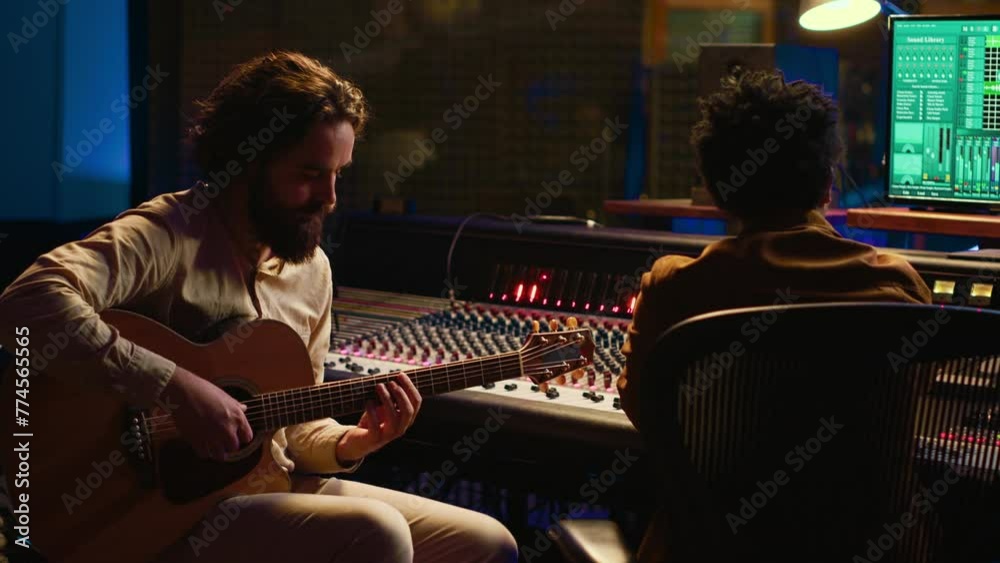 Creative artist singing on his electro acoustic guitar to record new track, producing music in control room at professional studio. Skilled musician composer creating new songs for an album. Camera A.