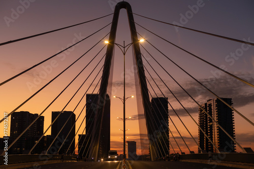 Sunrise on modern symmetrical suspension bridge with illumination, and road traffic. New high-rise buildings fill the background.
