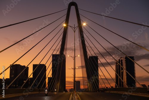 Sunrise on modern symmetrical suspension bridge with illumination, and motorcycle traffic. New high-rise buildings fill the background.
