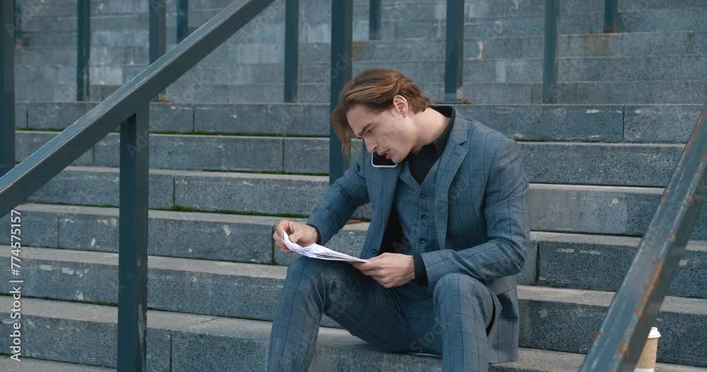 Businessman sits on steps engrossed in work report energetically conversing on phone Work report in hand he delves into strategies manifesting work dedication. Work calls echo his commitment.