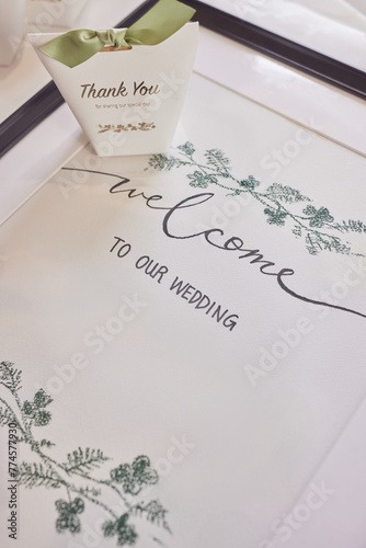 A white wedding welcome sign adorned with green floral designs, accompanied by a thank you note. photo