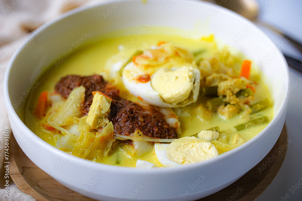 A bowl of “Lontong” or rice cake and coconut gravy with tumeric, chilli, and vegetables with some shadow and selective focus. A bowl of “Lontong” or rice cake and coconut gravy with tumeric, chilli, and vegetables with some shadow and selective focus.