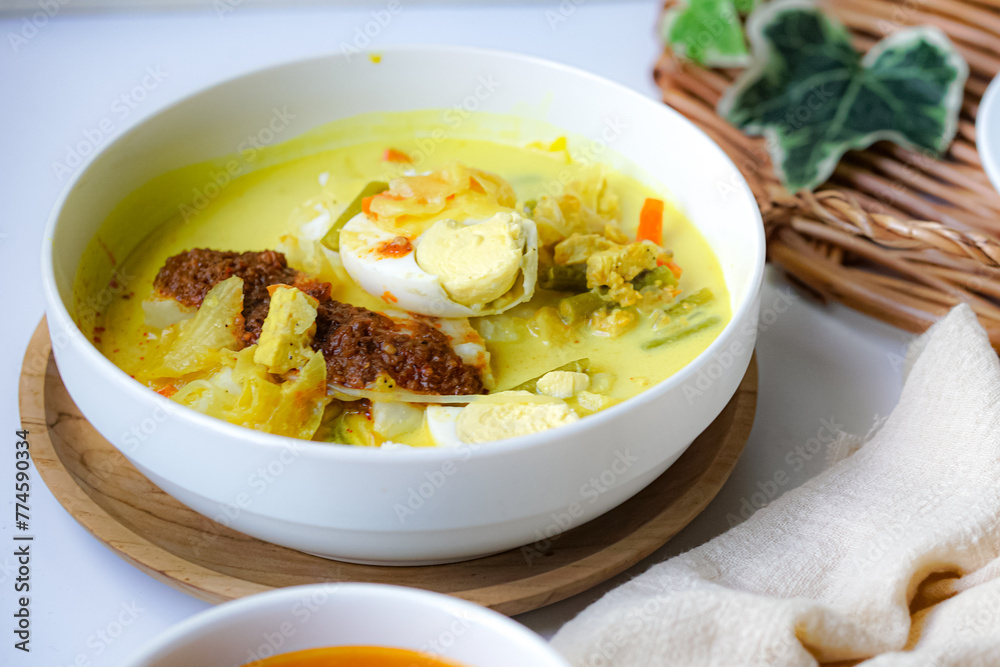 A bowl of “Lontong” or rice cake and coconut gravy with tumeric, chilli, and vegetables with some shadow and selective focus. A bowl of “Lontong” or rice cake and coconut gravy with tumeric, chilli, and vegetables with some shadow and selective focus.