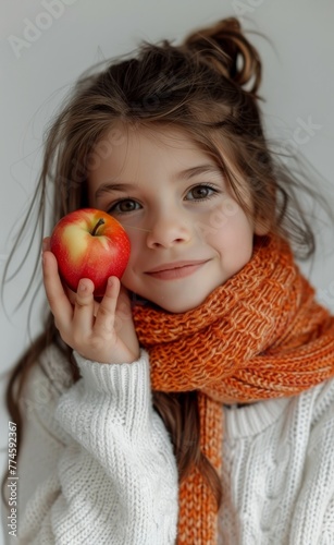 Smiling girl with apple in cozy knit scarf. Sweet smiling young girl holding an apple close to her face, surrounded by warm, orange knitwear