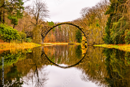 The Rakotzbrucke, also known as Devils Bridge, is reflected in calm waters on an overcast autumn day, surrounded by vibrant foliage. Germany © pyty