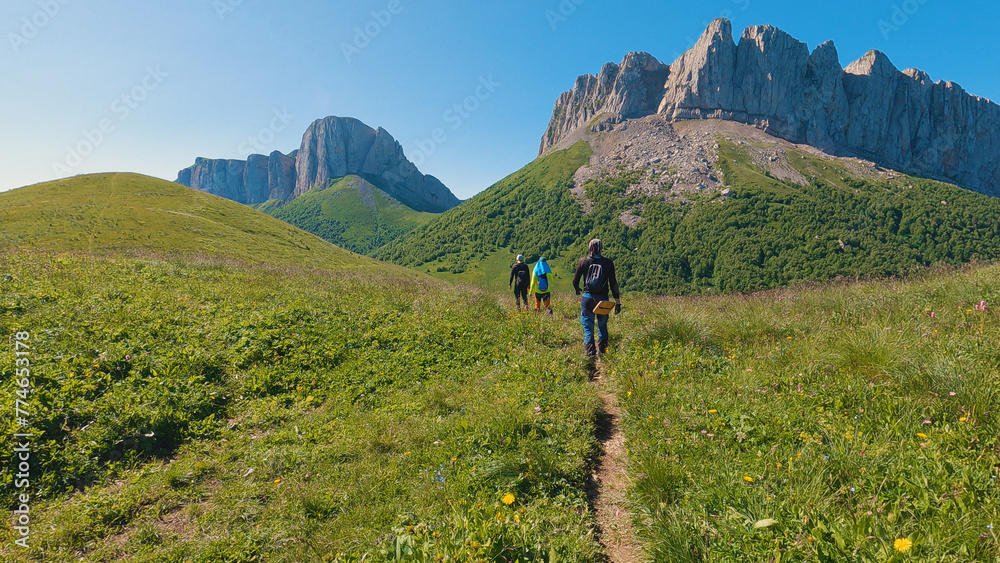 Fototapeta premium A group of hikers make their way along a narrow trail amidst lush greenery, heading for a rocky peak under a clear blue sky. Dressed in outdoor gear, they hike through the scenic mountain landscape.