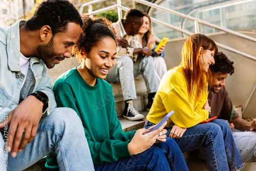 Group of young people using mobile phone sitting at staircase in the city. Millennial student friends having fun watching social media content on cell app. Technology lifestyle concept