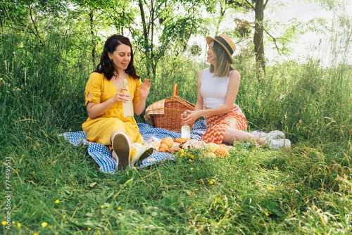 Pair of women sipping lemonade from bottles on blue picnic blanket. © phpetrunina14