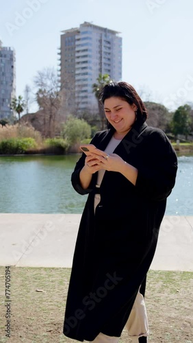 Curvy woman using phone while walking in a park. Person text messaging.