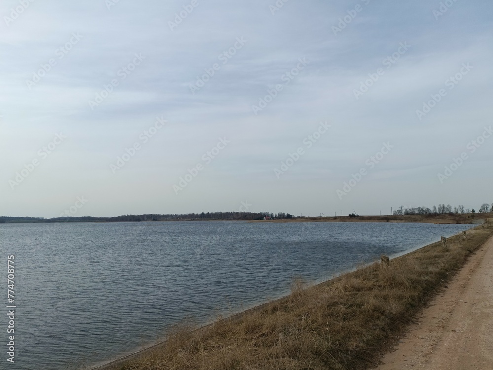 Gudeliai lake and lakeshore during sunny day. Lake with small waves. Lake shore with grass and reeds growing on it. Sunny day with white and gray clouds in blue sky. Nature. Gudeliu ezeras.