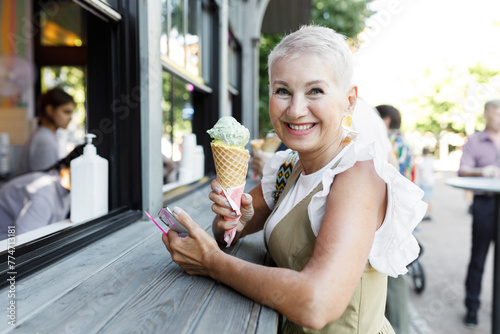 Side view of cheerful aged hipster female with short pink hair leaning on counter after buying waffle ice cream outdoor, looking at camera with candid childish smile during summer stroll in city