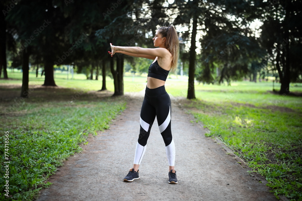 Sporty girl is doing exercises in park to warm up. Thanks to gymnastics in open air, volume lungs becomes larger. Girl for whole day you are charged with positive mood. Arm exercise with twist.