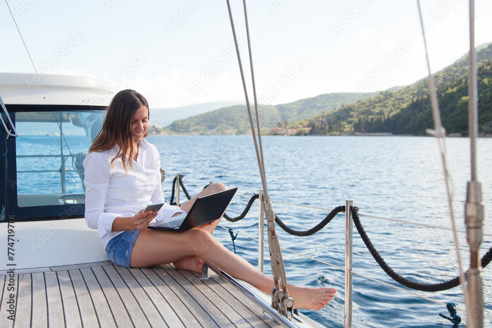 Happy woman working on yacht with laptop. Summer vacation on sailboat. Girl traveling and using computer, mobile phone, Internet. Freelancer office workplace outdoors. Successful business lifestyle