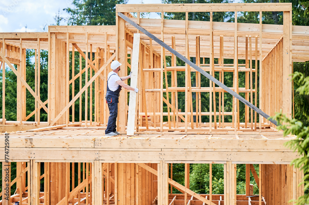 Carpenter constructs wooden-framed house. Man holds large plank in his hands while dressed in work clothes and helmet. Concept of modern ecological construction. Carpenter constructs wooden-framed house. Man holds large plank in his hands while dressed in work clothes and helmet. Concept of modern ecological construction.