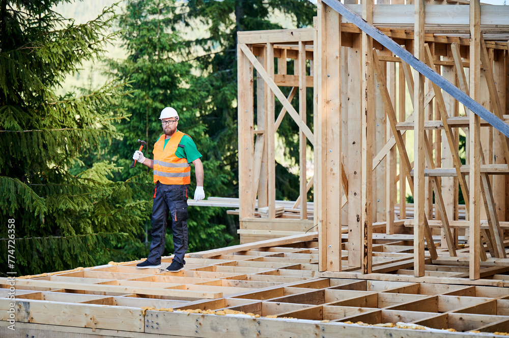 Carpenter constructing wooden frame two-story house near the forest. Bearded man holding hammer, dressed in protective helmet and orange safety vest. Concept of ecological modern construction. Carpenter constructing wooden frame two-story house near the forest. Bearded man holding hammer, dressed in protective helmet and orange safety vest. Concept of ecological modern construction.