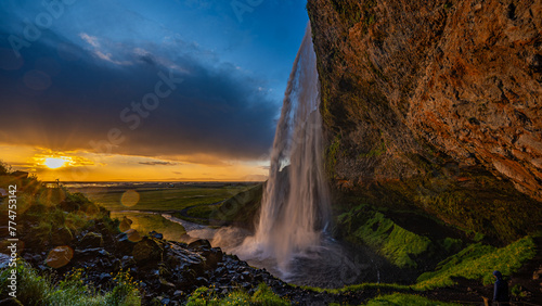 Die Mitternachtssonne scheint neben dem Wasserfall Seljalandsfoss  Weitwinkelaufnahme zeigt orange Sonne vor dunklen Wolken 