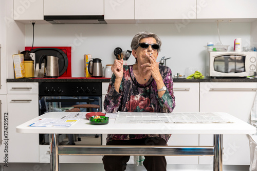 Old lady full of wrinkles smokes comfortably at home in her kitchen wearing black sunglasses. Funny situation