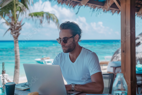 A young man in a T-shirt works on a laptop at a table on the beach in the summer. © SerPhoto