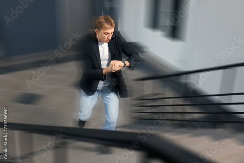 Being late. Young man checking time while running up stairs indoors. Motion blur effect