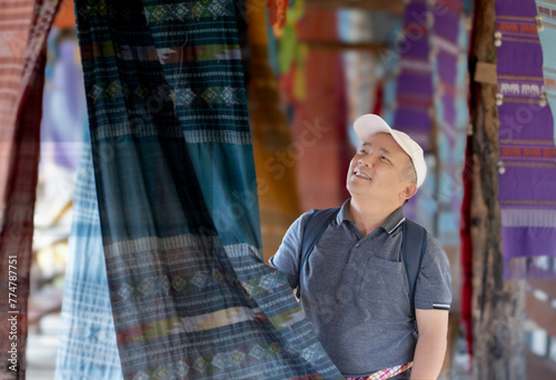 Asian middle-aged man in white cap and t-shirt holding local handmade woven fabric which hanging and showing for tourists in local fabric clothes shop, soft focus, happiness of people concept. photo
