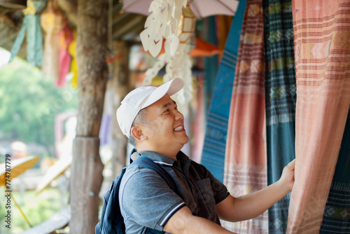 Asian middle-aged man in white cap and t-shirt holding local handmade woven fabric which hanging and showing for tourists in local fabric clothes shop, soft focus, happiness of people concept. photo