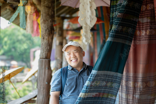 Asian middle-aged man in white cap and t-shirt  smiling  and holding woven fabric which hanging and showing for tourists in local fabric clothes shop, soft focus, happiness of people concept. photo