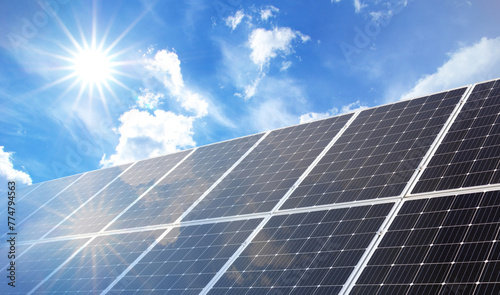 Solar Panels on Blue Sky with Sun Flares. Series of solar panels tilt upwards towards the clear blue sky, where the sun shines brightly casting a flare. Perfect day for harnessing solar energy.