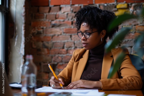 A businesswoman in an office, sitting at her desk and writing on paper with a pen, wearing a brown blazer over a black turtleneck sweater, a brick wall behind the woman, a computer screen