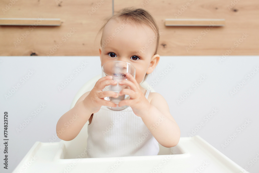 Sweet baby girl holding glass and drinking water. Happy baby sitting in a baby chair in the kitchen. Sweet baby girl holding glass and drinking water. Happy baby sitting in a baby chair in the kitchen.