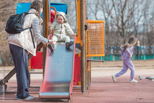 A beautiful, cheerful little boy of two years old walks with his mother on the playground in the spring. Mom rolls the baby down a slide in the park. The concept of parenthood, childhood and family.