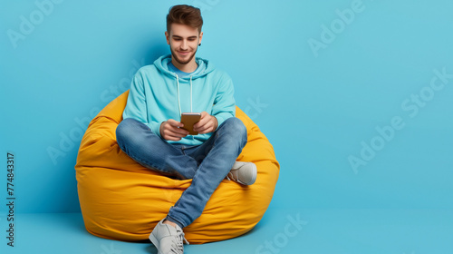 Photo of a smart, positive guy in a sweatshirt and denim pants, sitting on an ottoman and looking at a smartphone, on a blue color background. Copyspace.