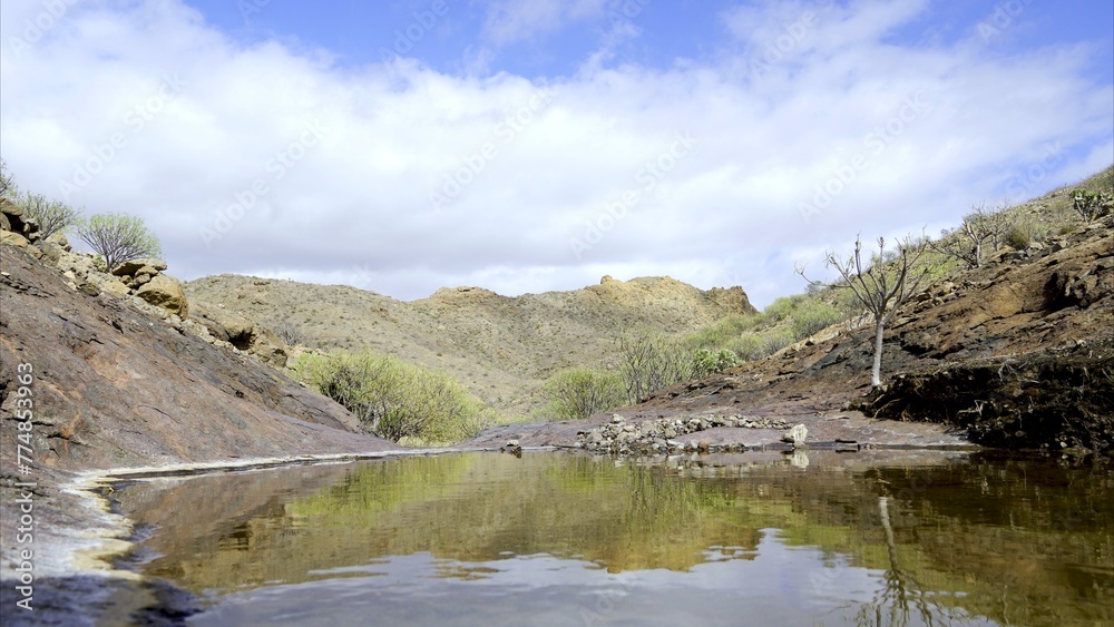 The banks of the stream are reflected in the still water with a cloudy sky and a barren mountain in the background The banks of the stream are reflected in the still water with a cloudy sky and a barren mountain in the background
