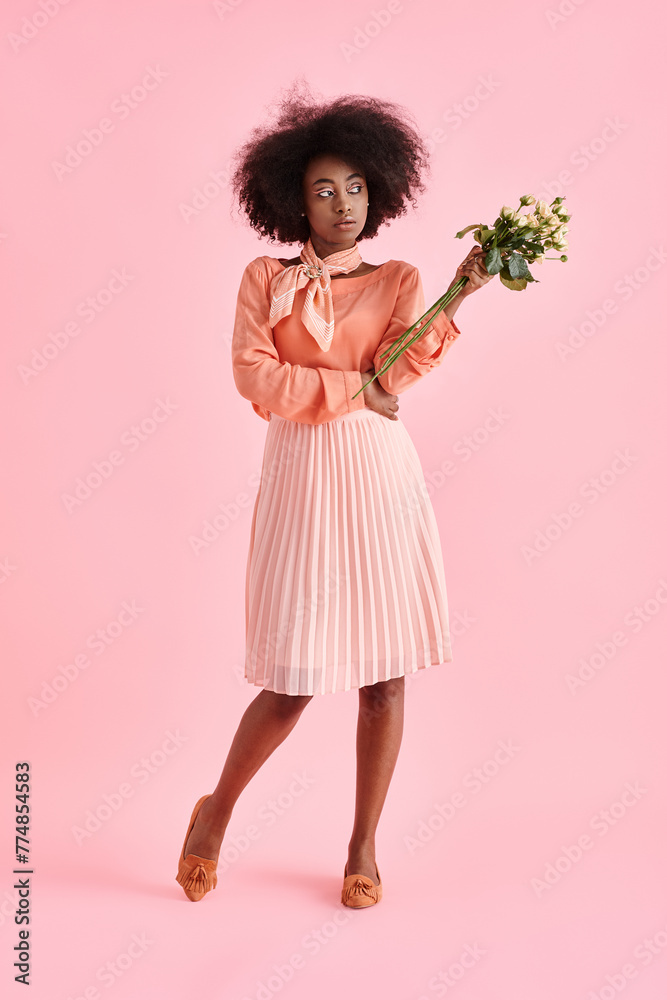 african american woman in peach fuzz outfit holding flowers and looking away on pink background
