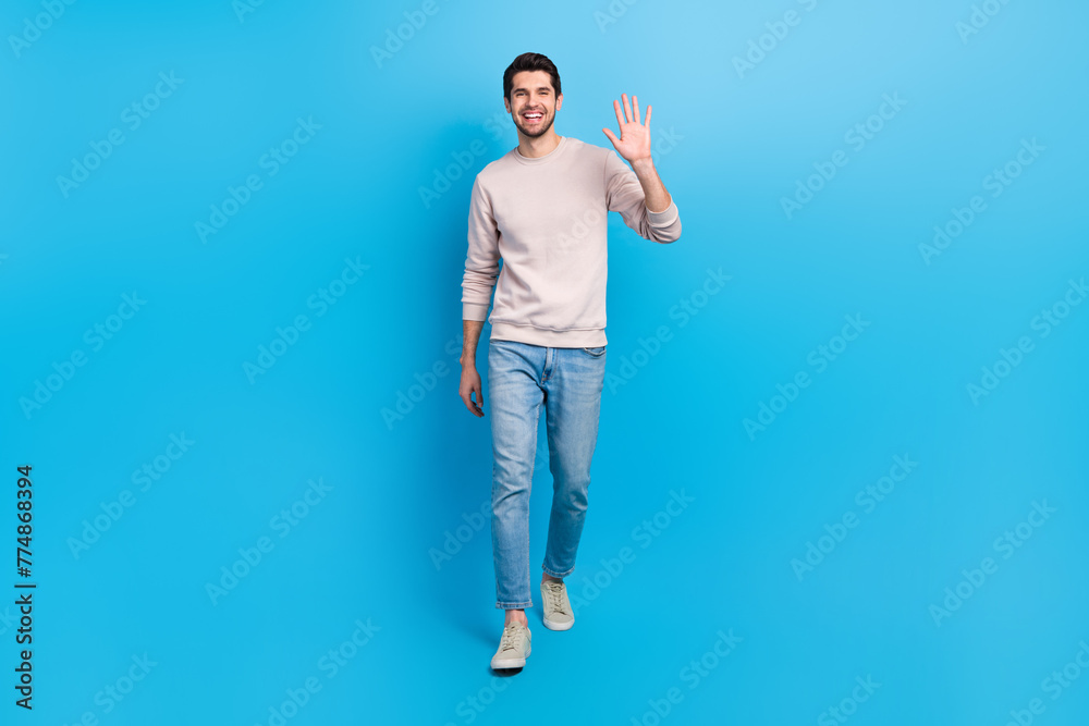 Full size photo of optimistic cheerful positive man wear sweatshirt denim pants waving palm say hello isolated on blue color background Full size photo of optimistic cheerful positive man wear sweatshirt denim pants waving palm say hello isolated on blue color background